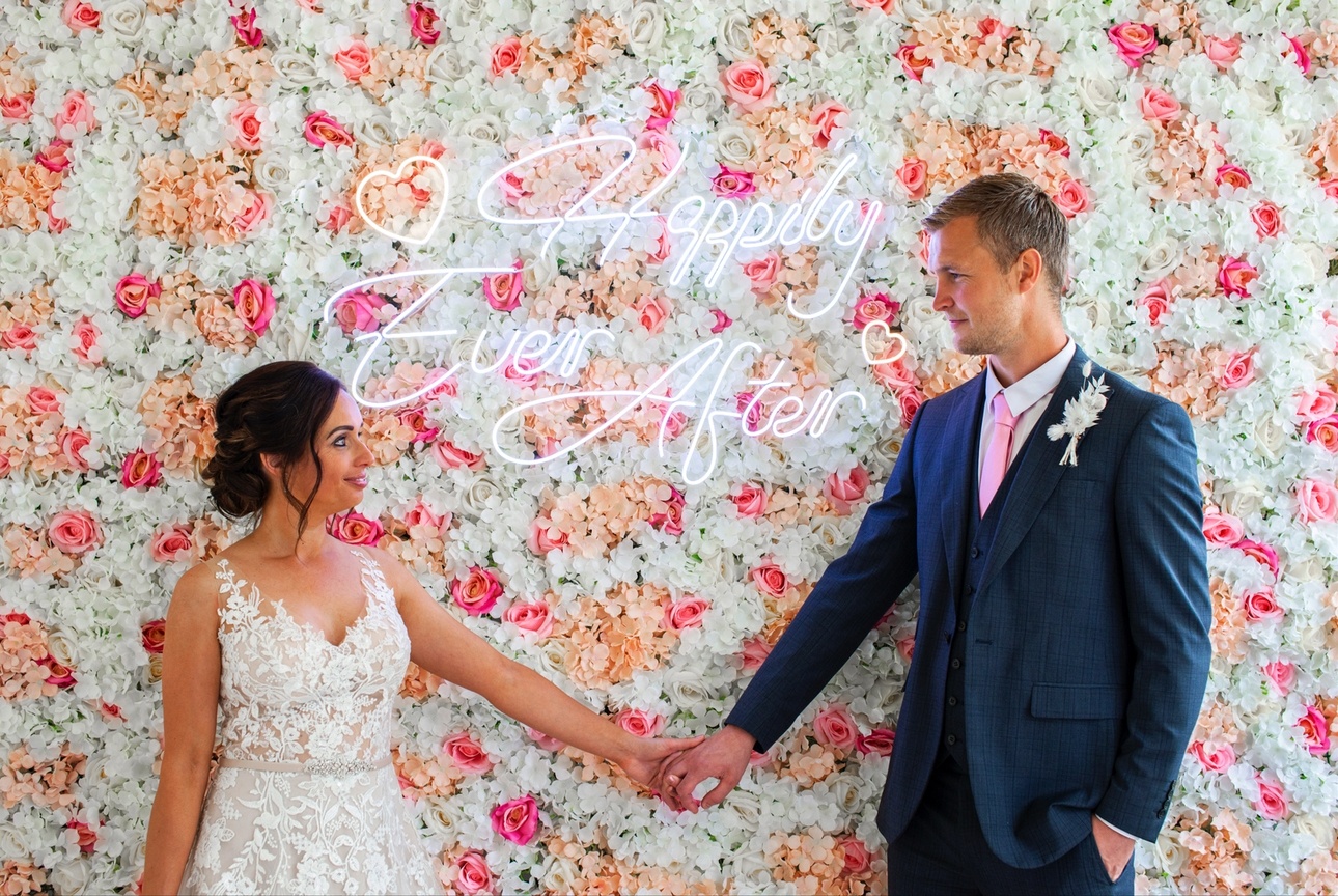 Bride and groom in front of a flower wall with neon sign saying Happily Ever After