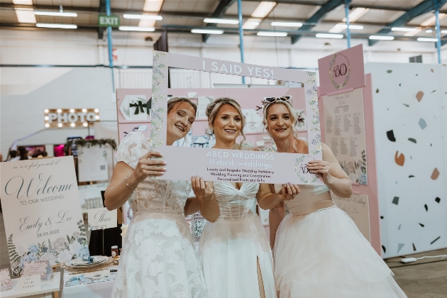 Three catwalk brides at The Big Southwest Wedding Fair hold up a selfie frame as they prepare to have their picture taken
